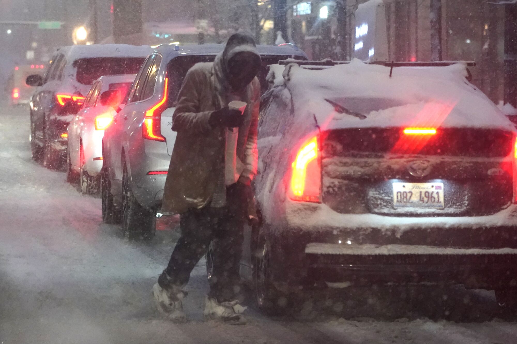 A person walks between cars as he navigates windy, falling snow and slushy street conditions