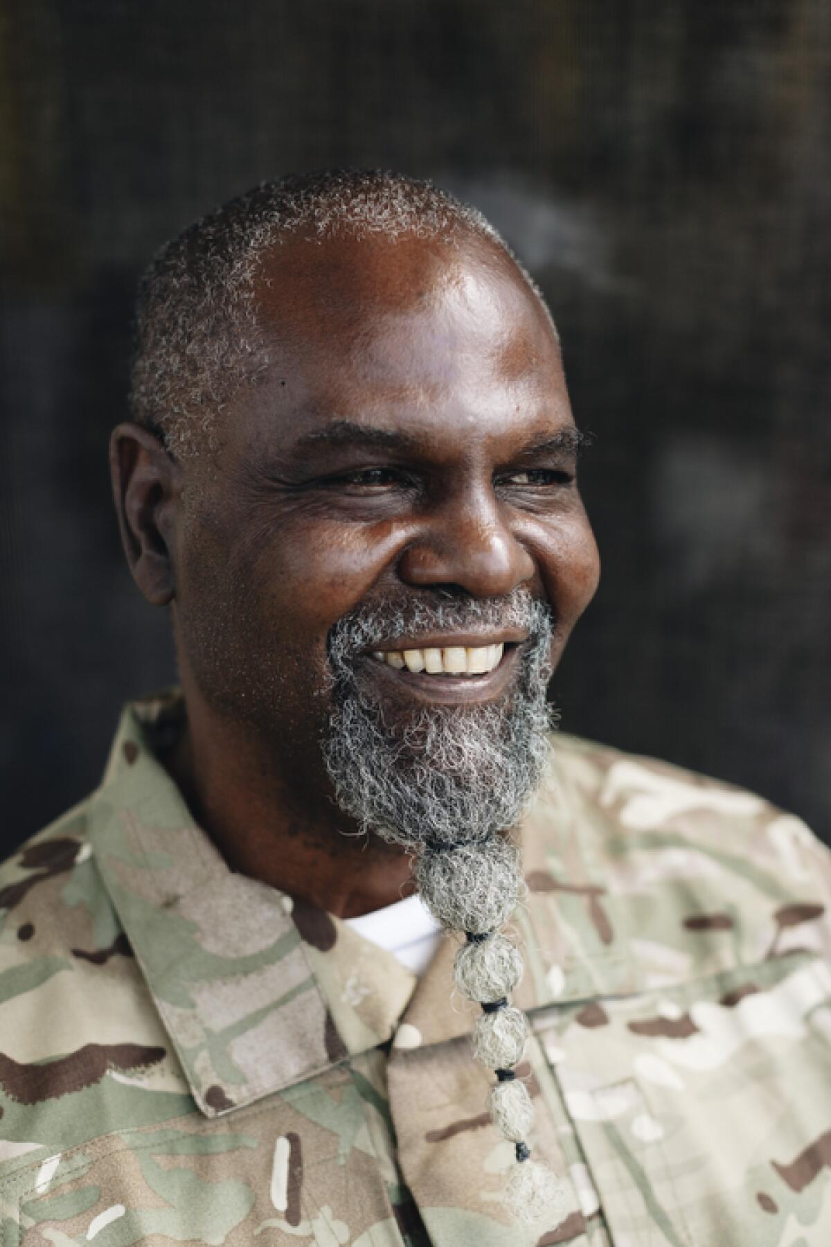 A vertical portrait shows a smiling Black man with a long beard that has been tied into a point.