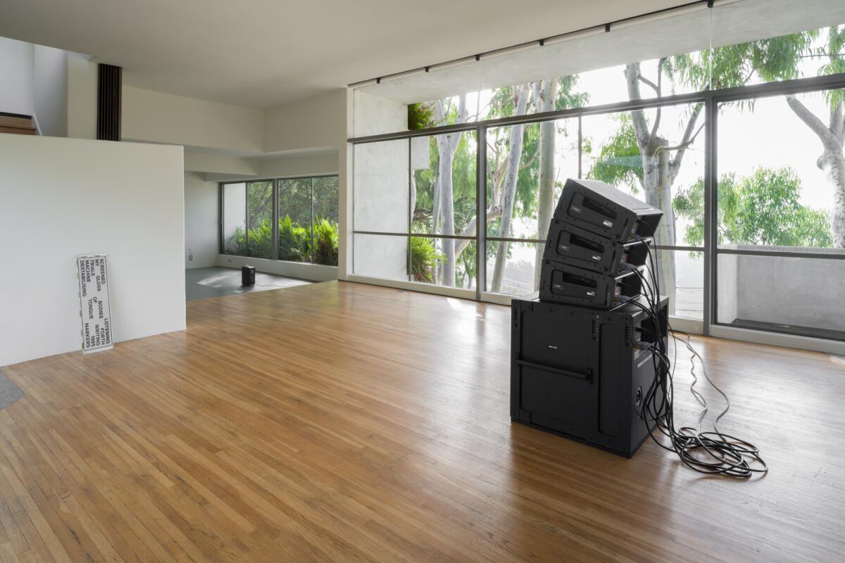 An empty room with broad windows features a stack of speakers and a text display element leaning against one wall.