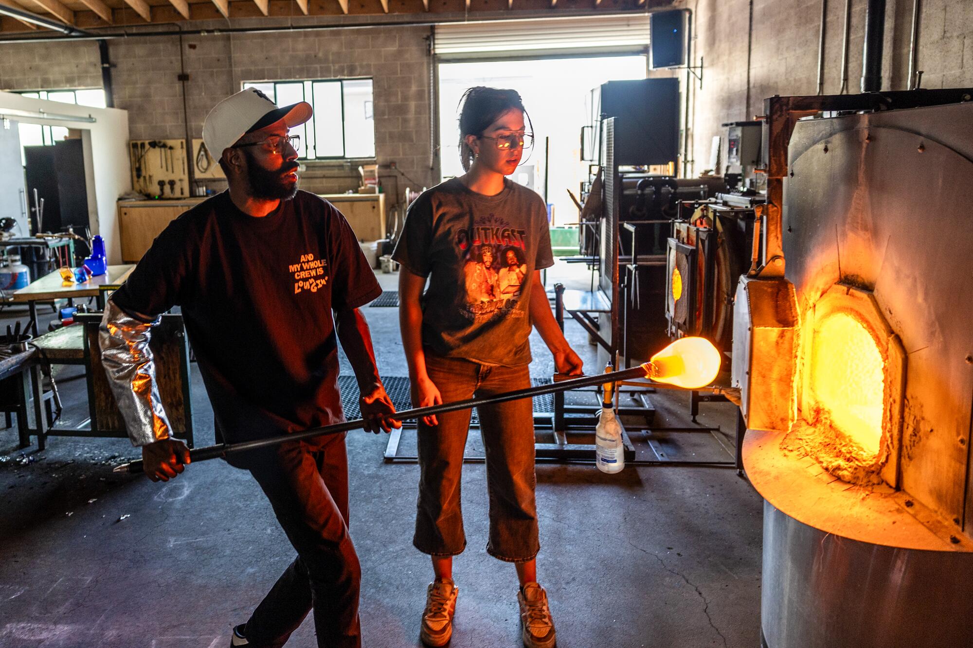 Assistant glassblower Sara Roller watches as glassblower Cedric Mitchell removes glass from a furnace.