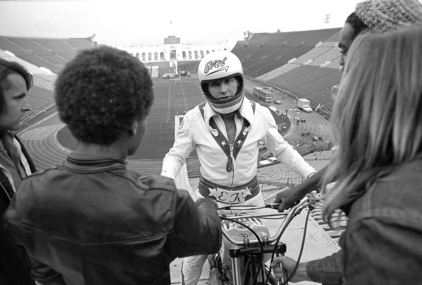 A man on a motorcycle in a helmet that says "Evel," with an empty stadium in the background.