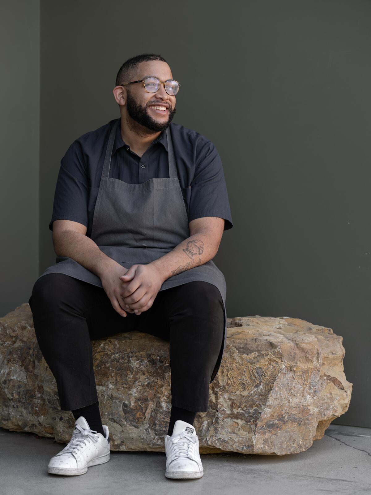 Josh Ulmer, a man in a chef's apron and glasses, sits on a large rock.