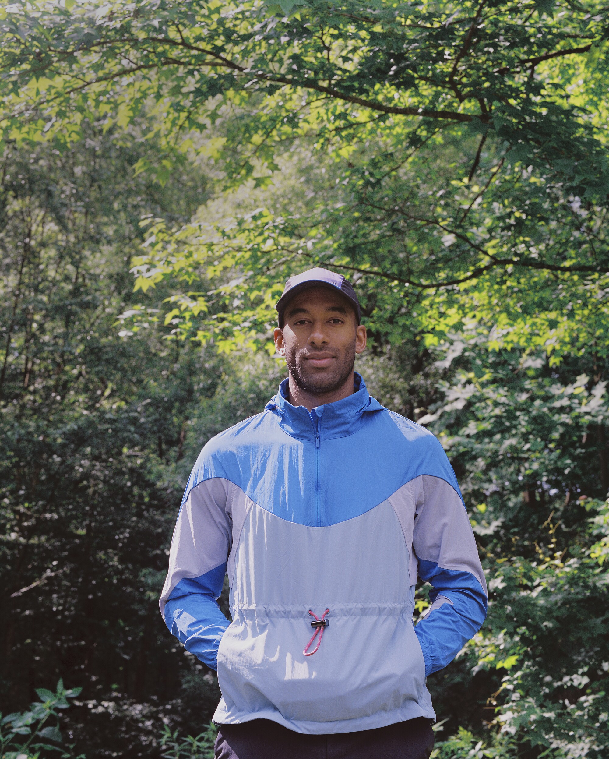 A man wearing a hat and blue windbreaker poses in front of trees.