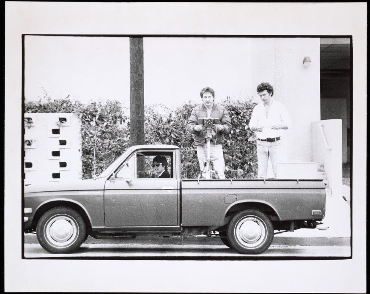 A black and white photo shows Danny Kwan at the wheel as Ed Ruscha and Bryan Heath stand in the bed of Ruscha's truck