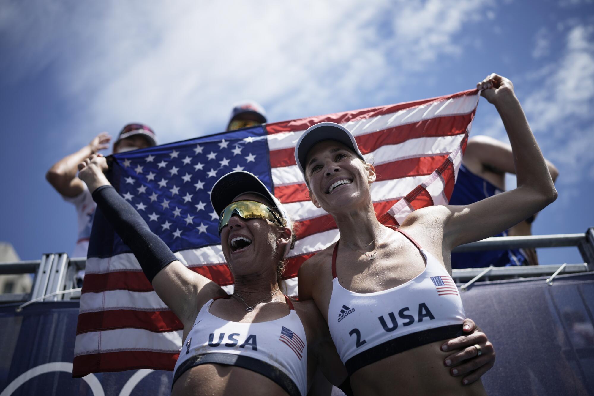 April Ross, left, and teammate Alix Klineman wearing USA jerseys as they hold up an American flag and celebrate.