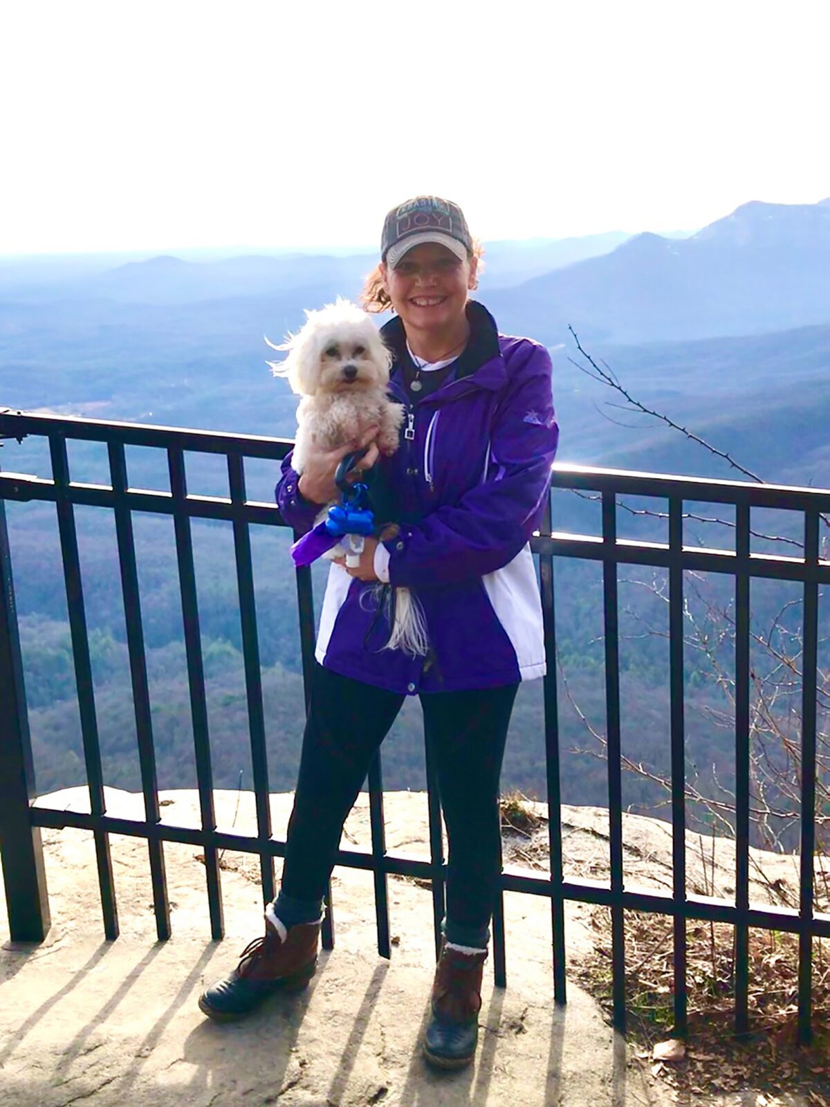 A woman holds a small white dog and poses for a photo in front of a mountain landscape.