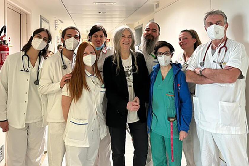 A woman in a black outfit surrounded by eight people in scrubs and white medical gear standing in a hospital hallway