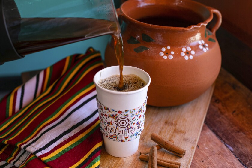 A pitcher pouring coffee into a white cup with a colorful wrap that says Azucanela, next to a Mexican red clay pot.