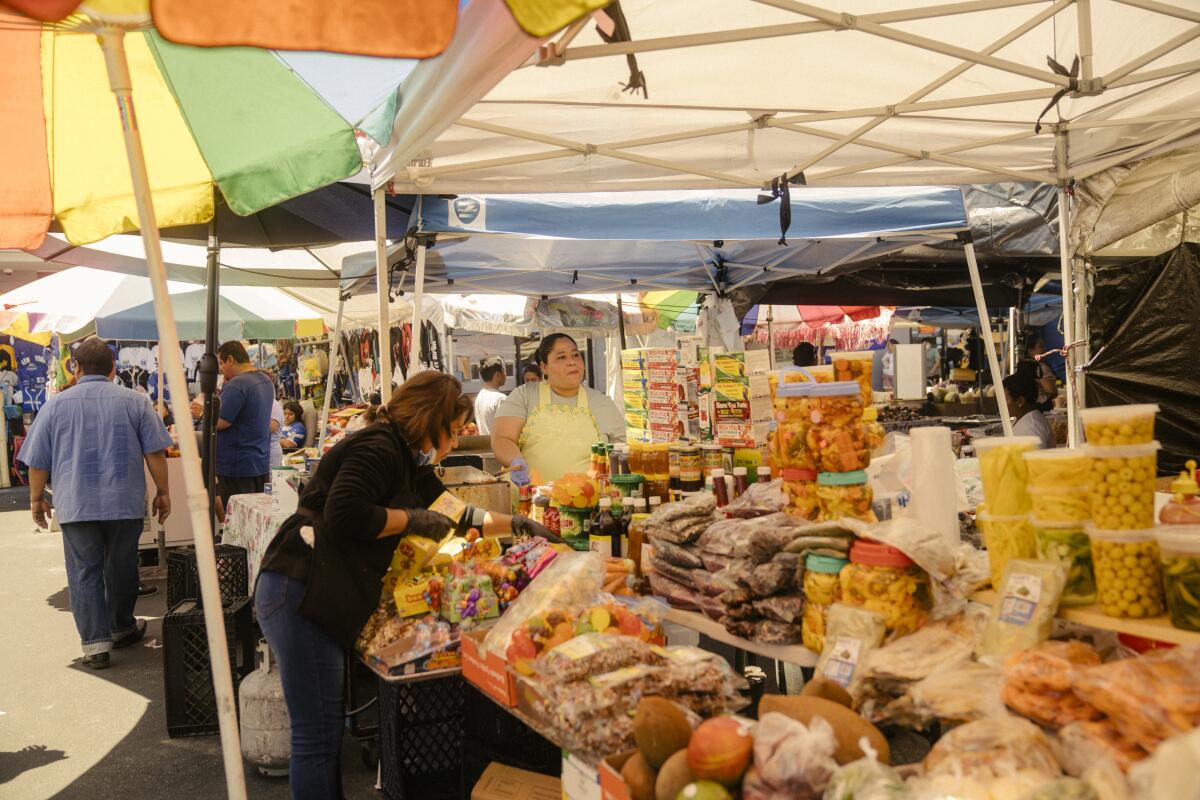 People stand alongside shelves full of produce and goods under an open-sided tent.