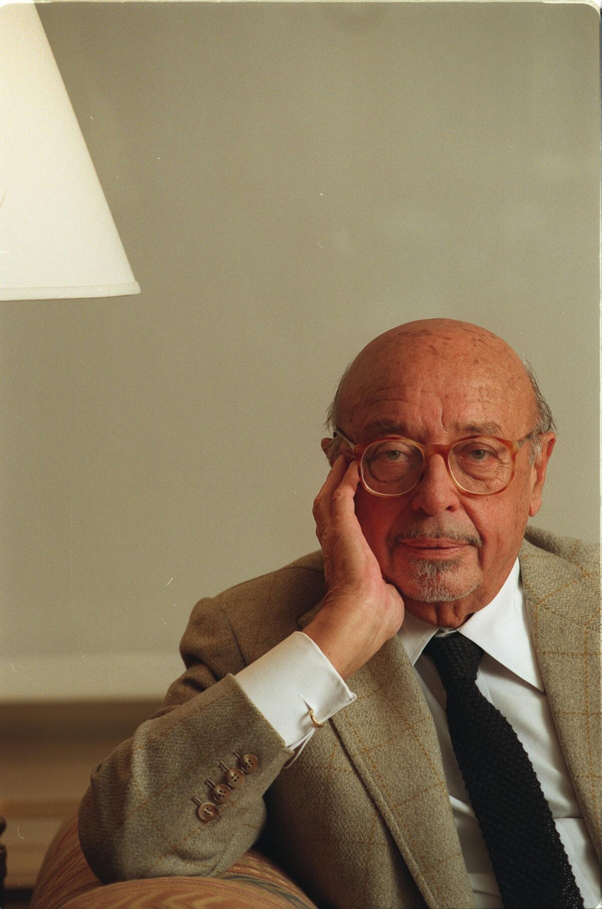 Man wearing a suit jacket and brown round glasses sits in a chair with his right hand against his face  