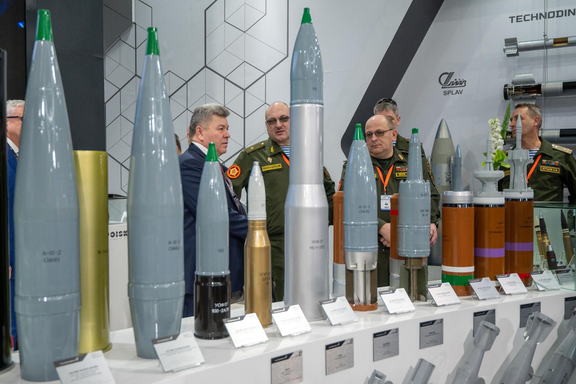 Men in suits and military uniform view a row of various projectiles on display 
