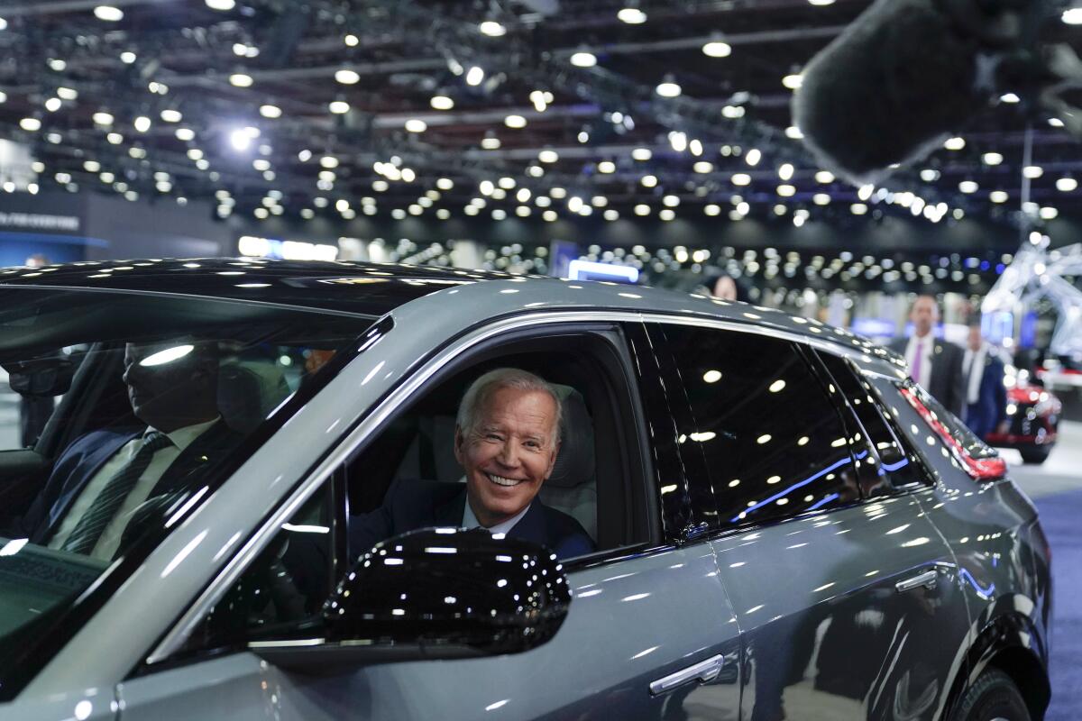 A smiling man in a dark suit in the driver's seat of a shiny gray vehicle