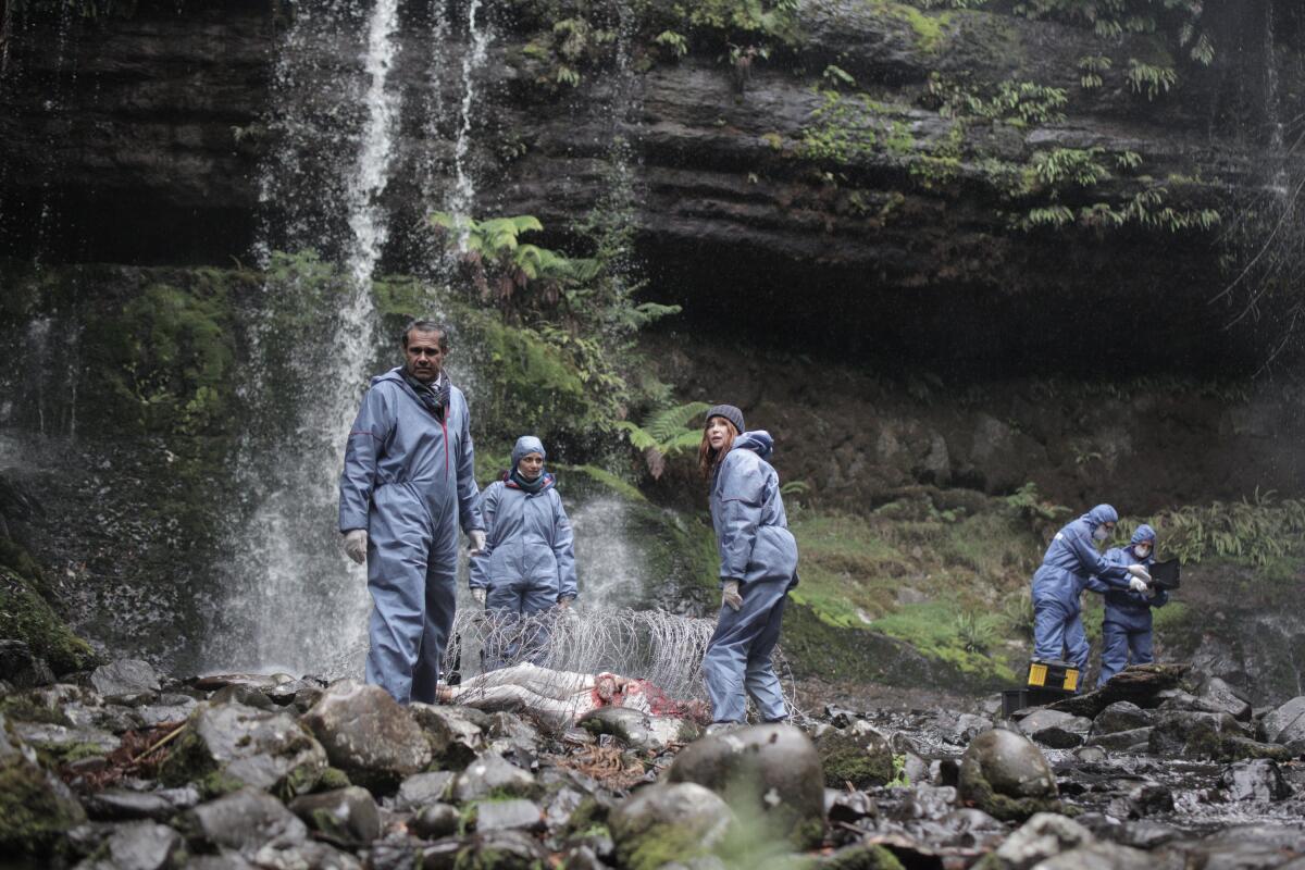 A group of police conduct forensic work on a corpse at the foot of a waterfall.