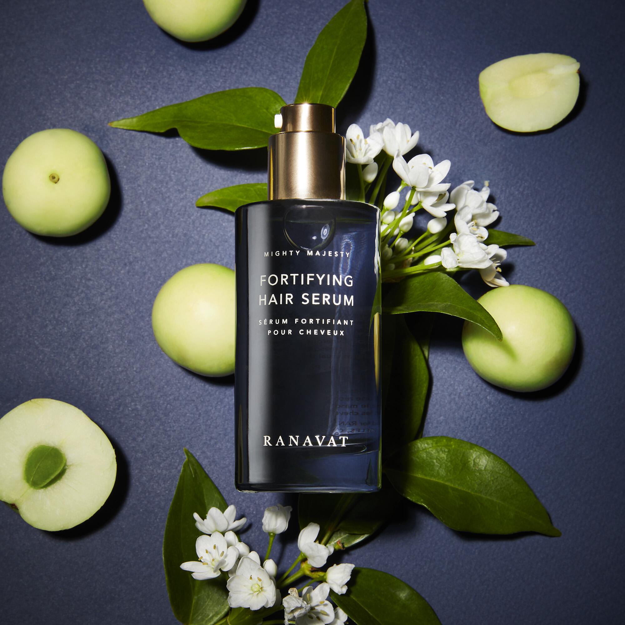 a blue Ranavat hair serum bottle lying on top of small white flowers and leaves, surrounded by green fruit