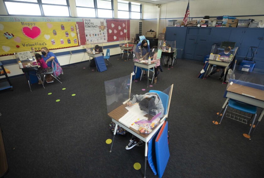 Students sit at spaced-out desks with plastic barriers