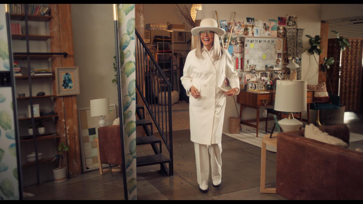 Diane Keaton, in white hat, trench coat and pants slacks, stands laughing in the middle of a living room.