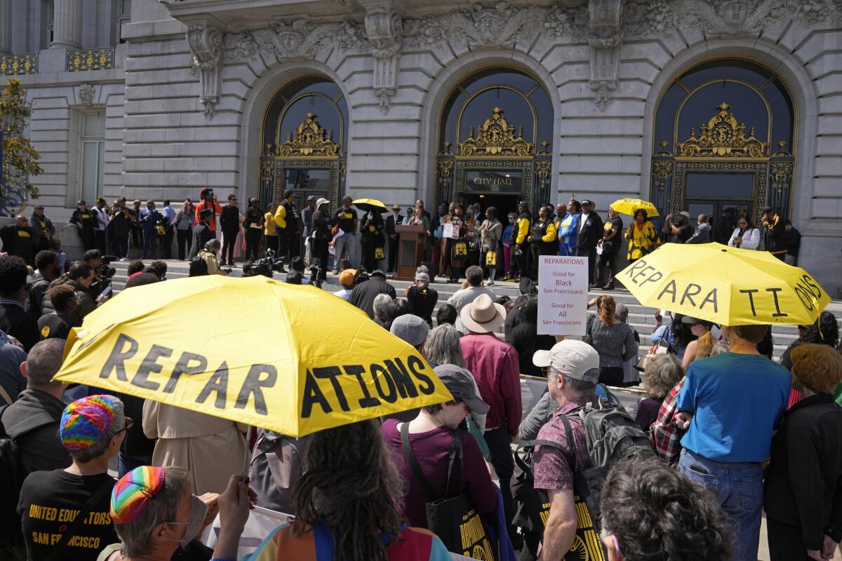 People at a rally outside San Francisco City Hall stand under yellow umbrellas with the word Reparations written on them