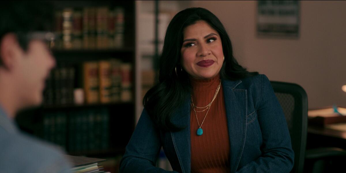 A woman with black hair wearing a blue suit and sitting at a desk