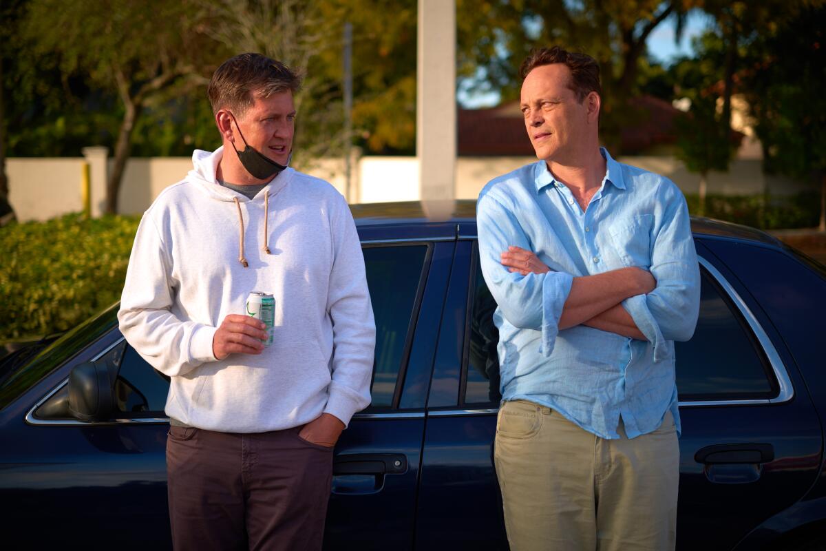 A man in a white hoodie leans against a car next to a man in blue shirt and khaki pants.