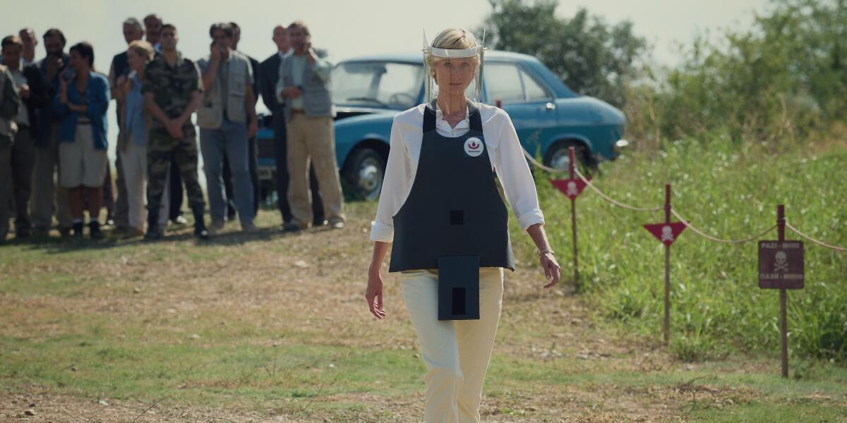Elizabeth Debicki as Diana in a white shirt, khakis, vest and face shield walking through a landmine field.