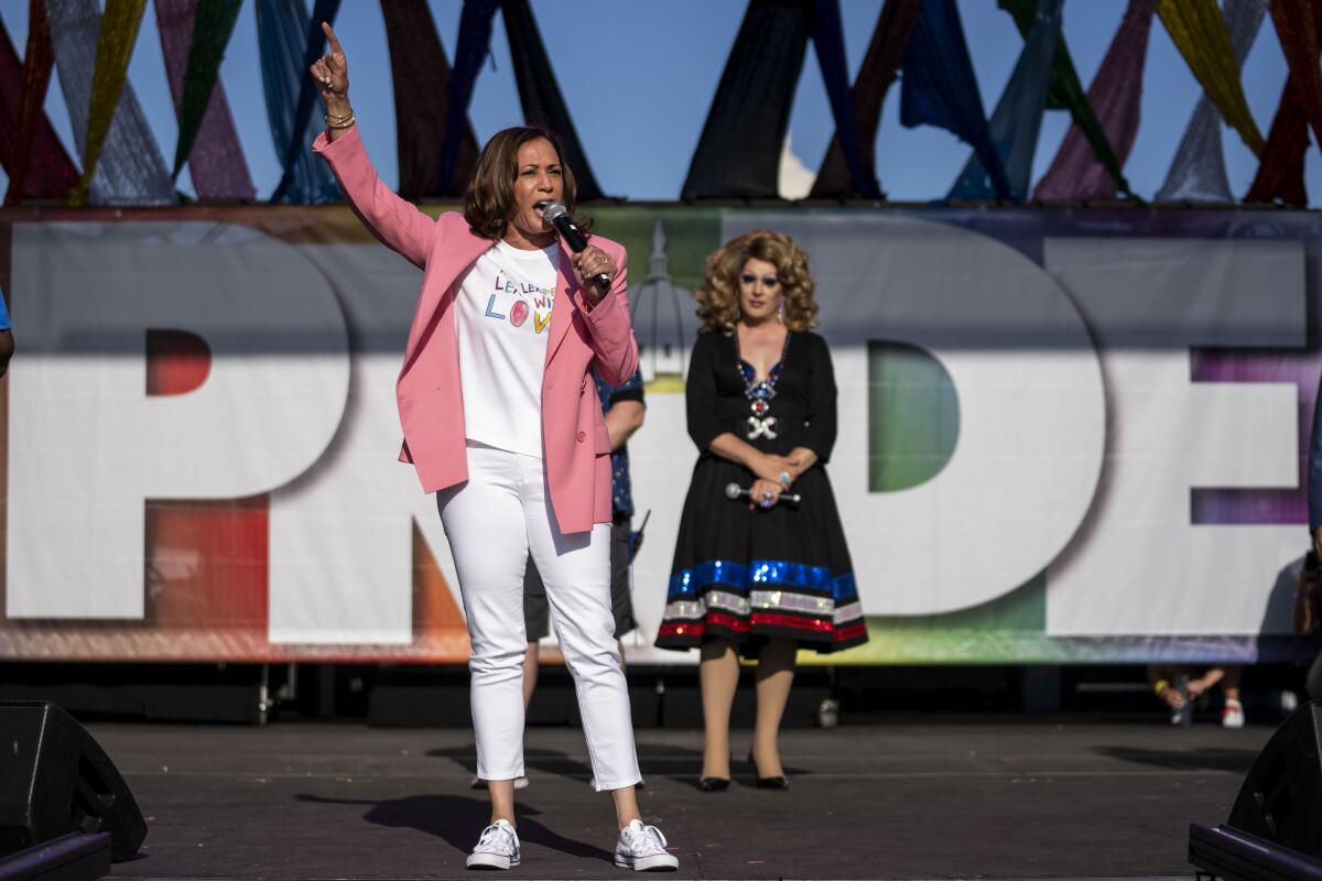 A woman in a pink jacket, white T-shirt and pants raises one hand as she speaks into a microphone.