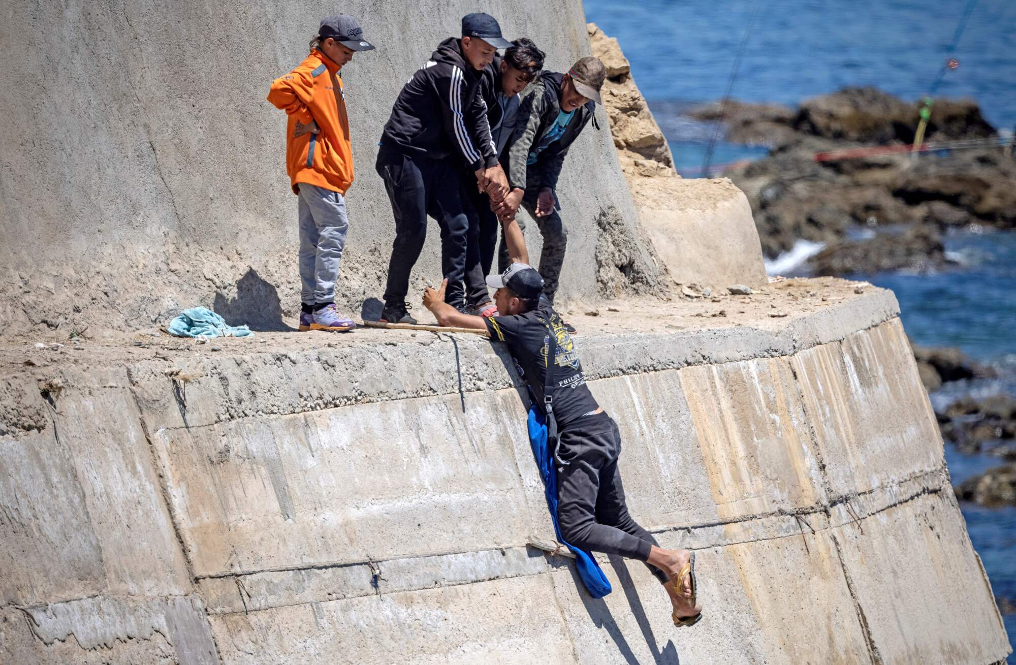 Four boys reach down to help pull up a fifth while climbing a concrete sea wall