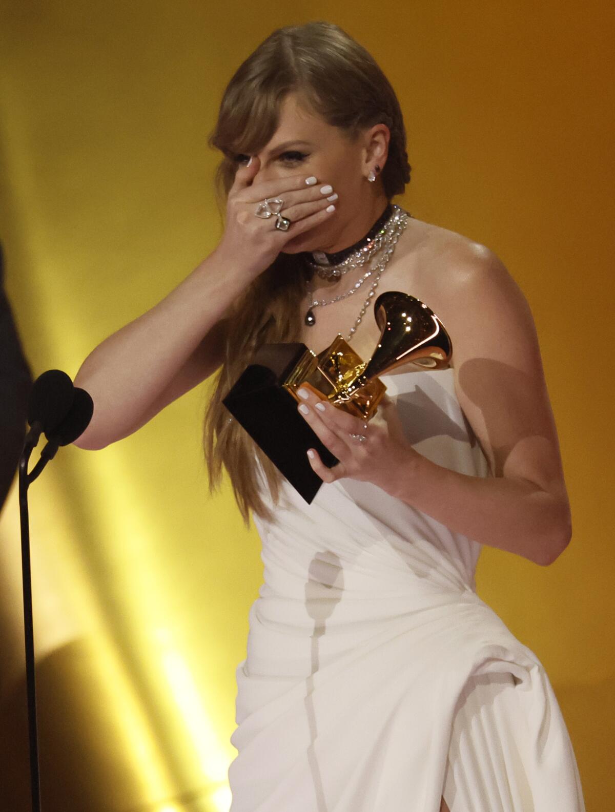 Taylor Swift holds her hand over her face in surprise and holds a Grammy in her other hand 