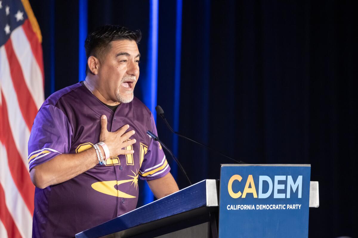 A man with dark hair, holding a hand to his chest while speaking at a lectern with the word CADEM
