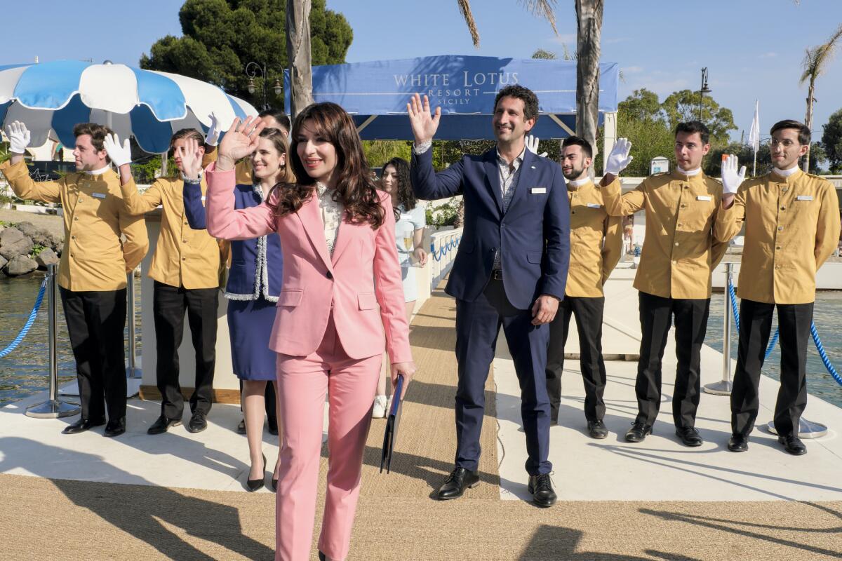 A woman in a pink pantsuit, flanked by a man in a blue suit and a row of men in ochre suits, waving from a dock.