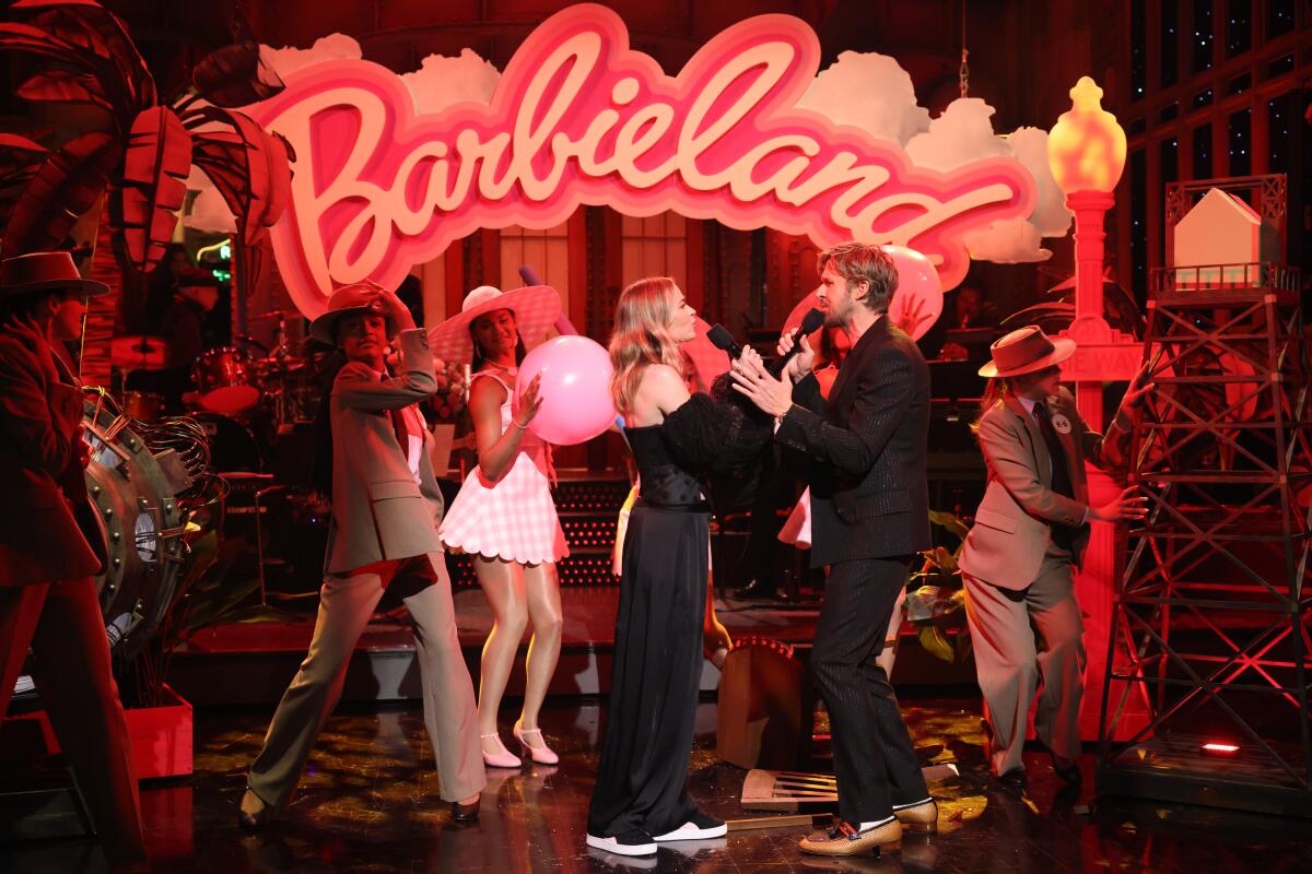 A man and a woman wearing black hold microphones and sing facing each other under a Barbieland sign