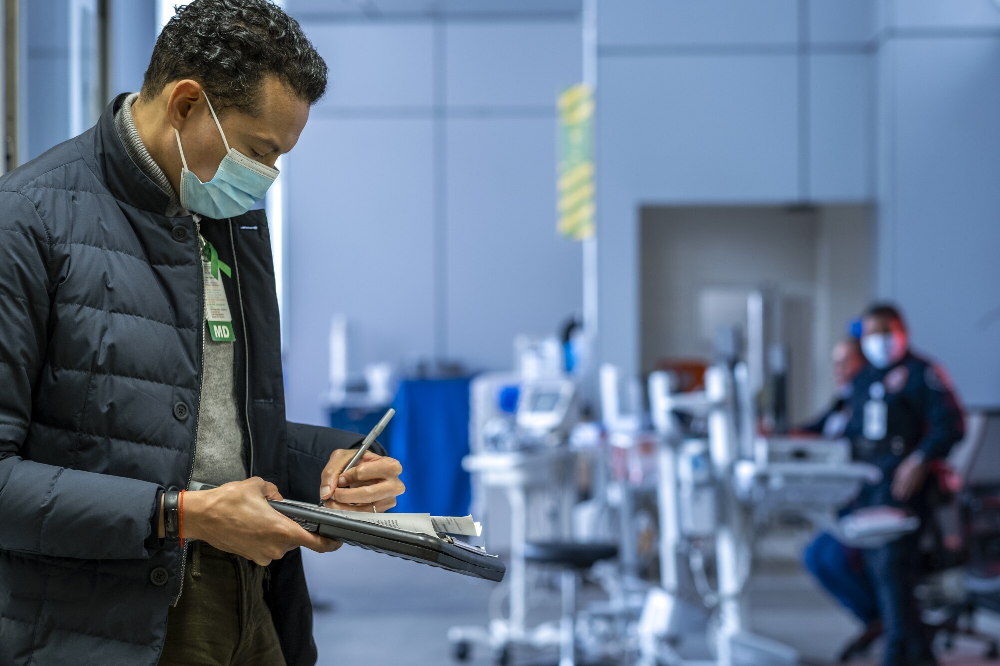 A masked doctor stands in the ER taking notes.