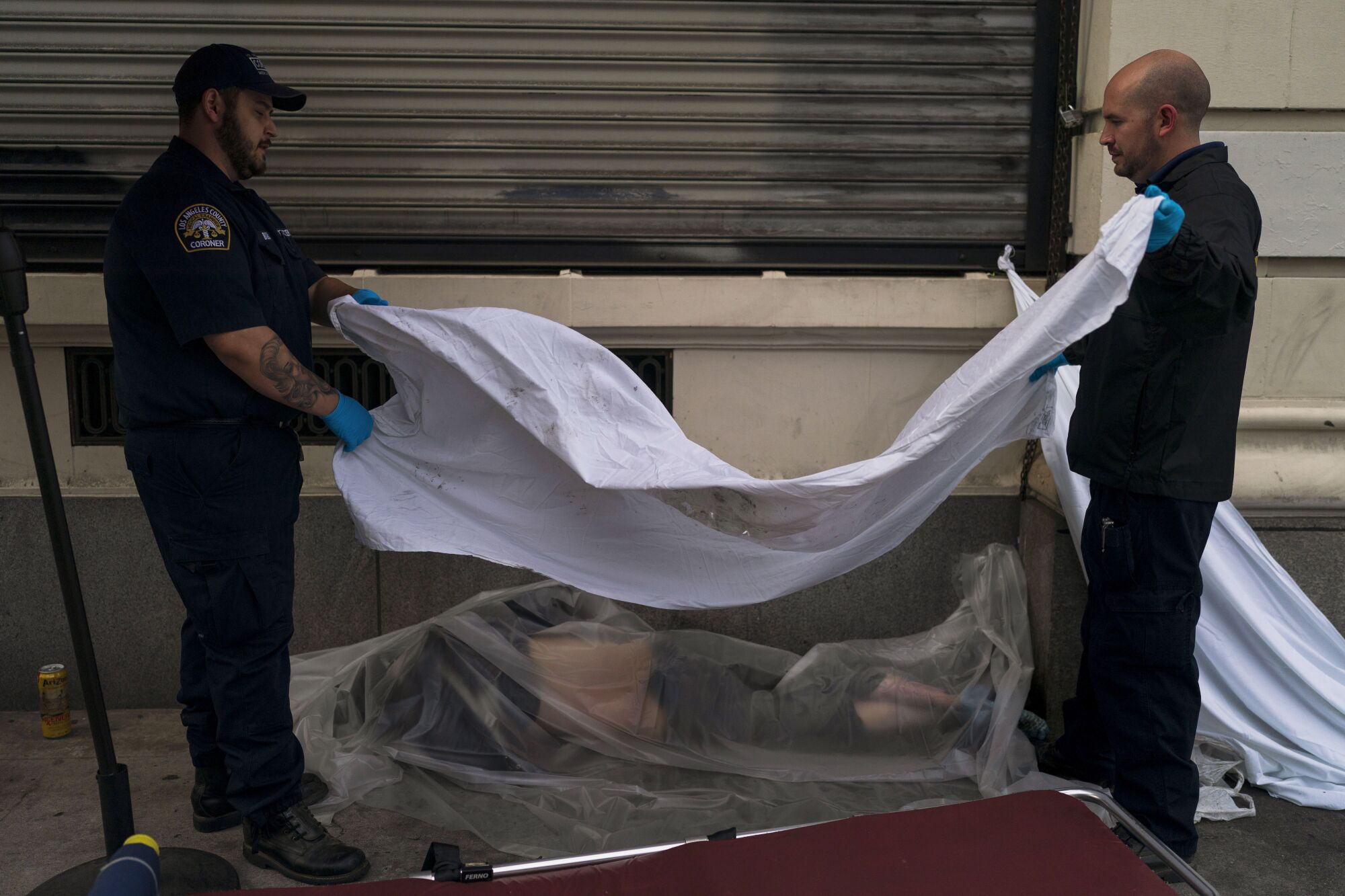 Zwei Männer benutzen ein weißes Laken, um den Körper eines Obdachlosen zuzudecken, der tot auf einem Bürgersteig in LA gefunden wurde.