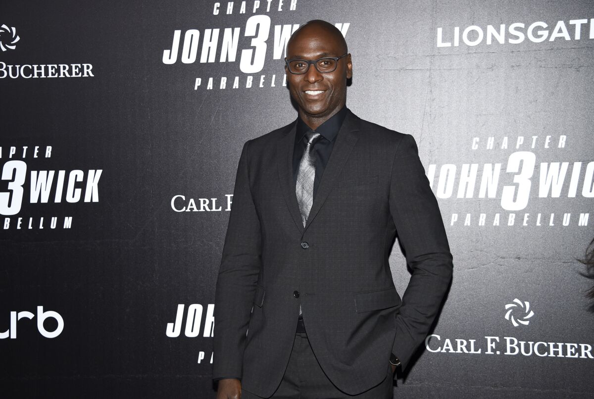 A bald, smiling actor wearing a dark suit stands with a hand in one pocket in front of a 'John Wick: Chapter 3' backdrop