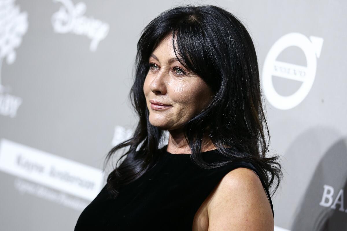 A side view of Shannen Doherty dressed in black and posing in front of a backdrop