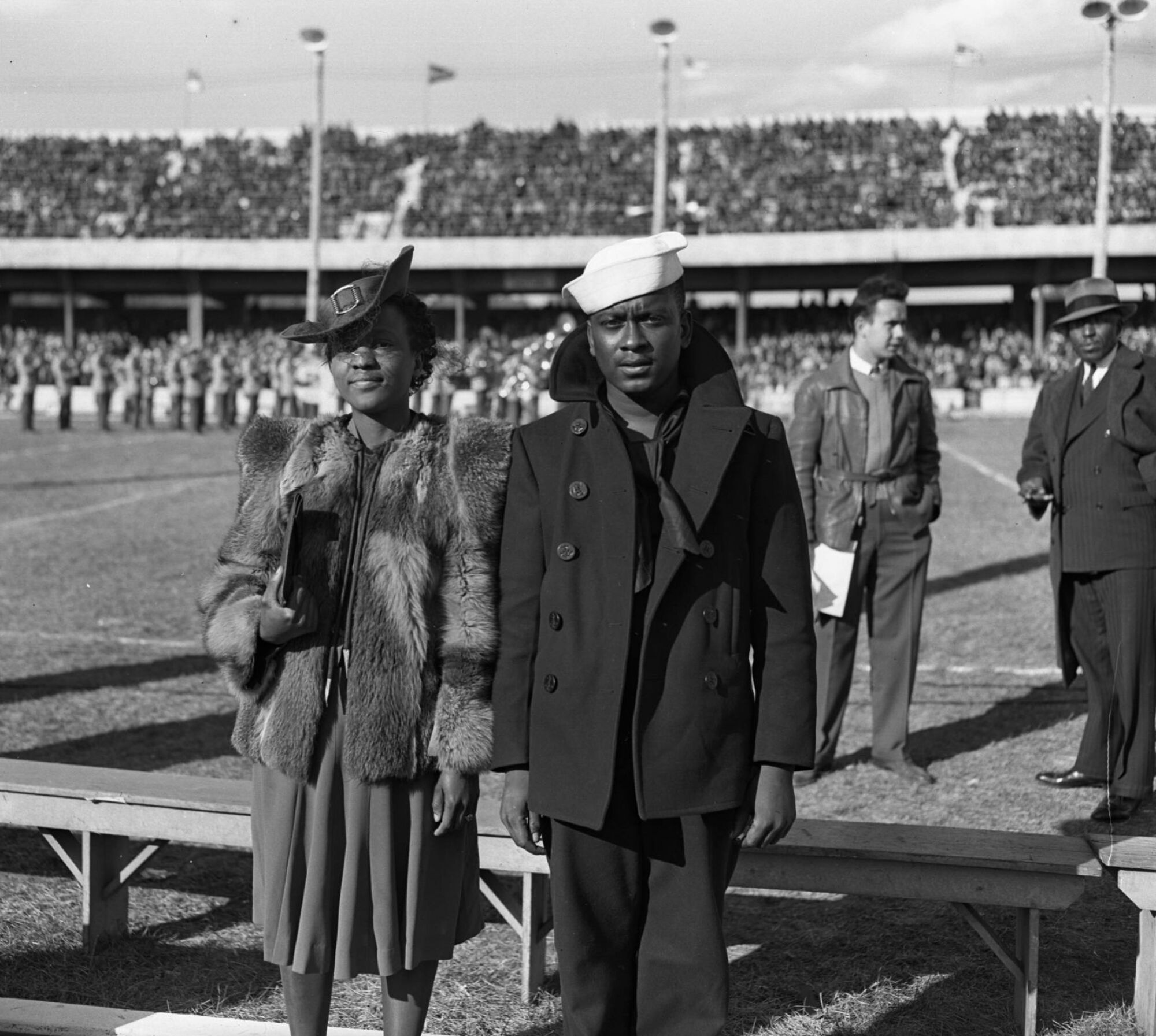 A man in a Navy uniform and coat stands next to a woman in a dress and coat at a football game.