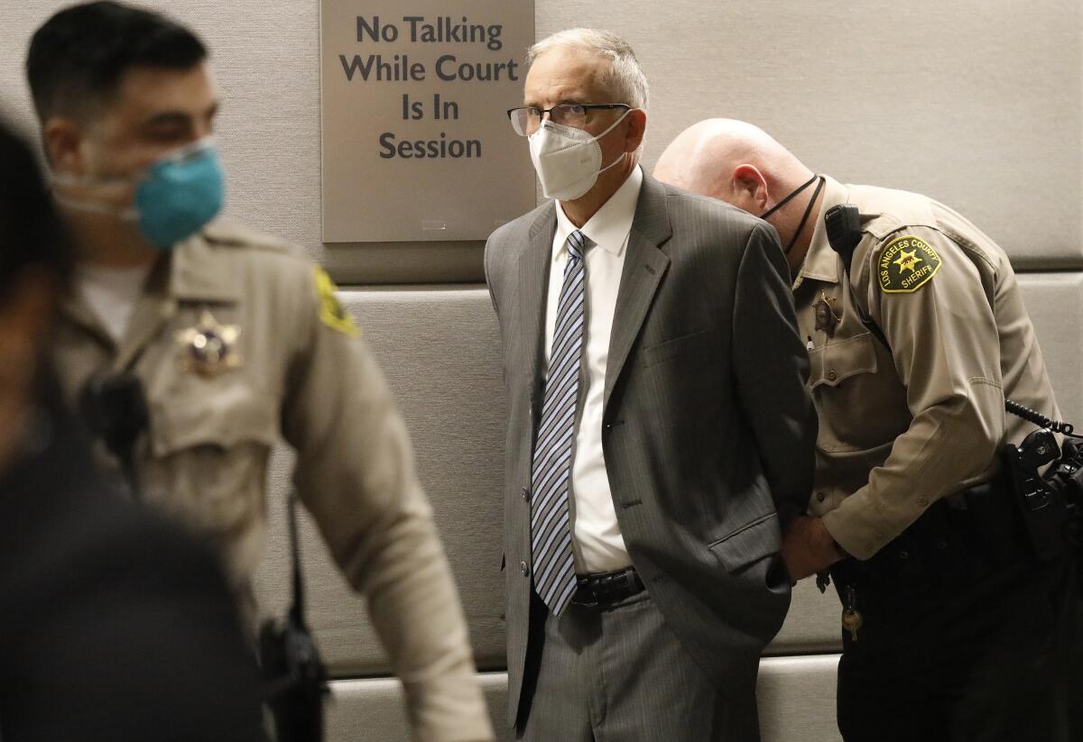 A man with short gray hair, glasses and face mask, and dressed in a suit, stands in a courtroom.