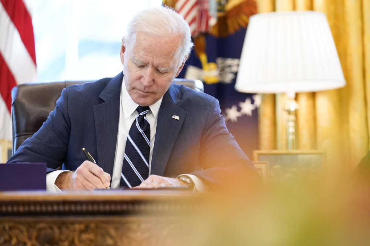 President Biden signing documents while sitting at his desk in the Oval Office. 