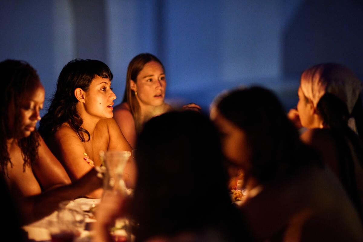 Naked people sit at a long dinner table lit by candles.