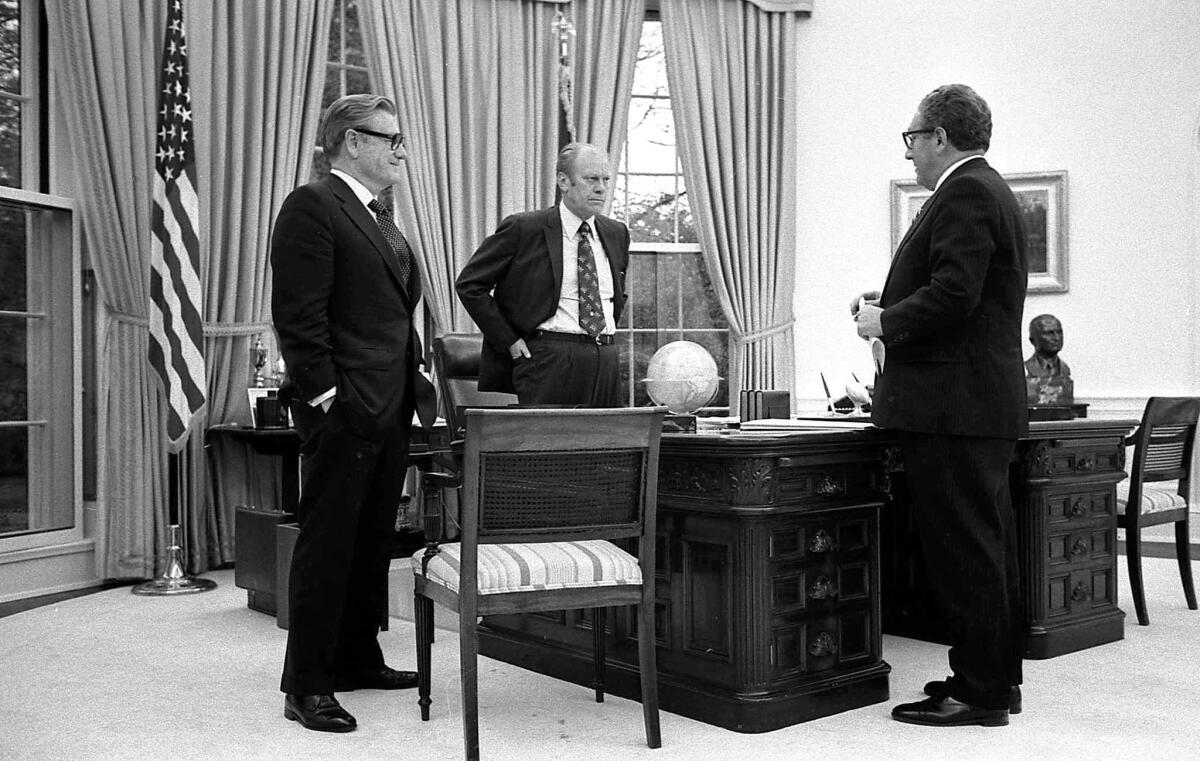 Three men in dark suits stand around a large desk