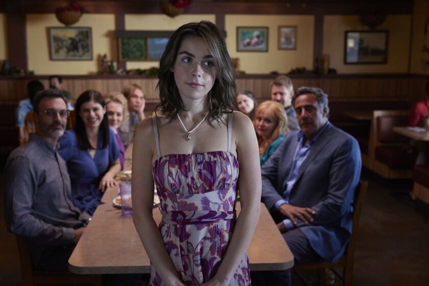A young woman standing in front of a table in a restaurant, where her family is seated in the movie "Wildflower."