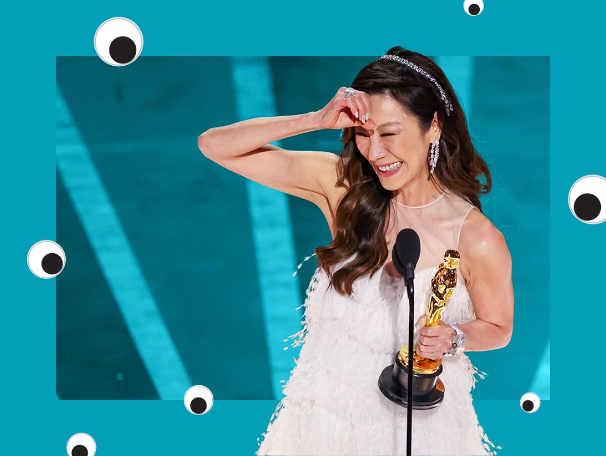 An Asian woman gleefully holds an Oscar in one hand and makes a circle gesture on her forehead with the other.