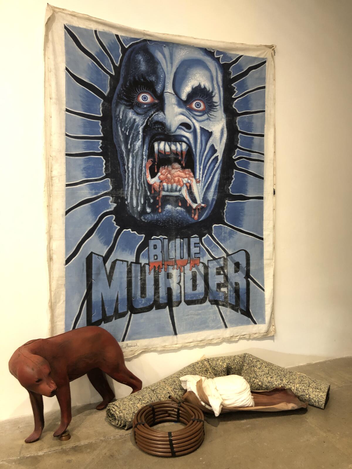 A sculpture of an animal and other objects sit before a billboard for "Blue Murder" showing a figure with vampiric teeth