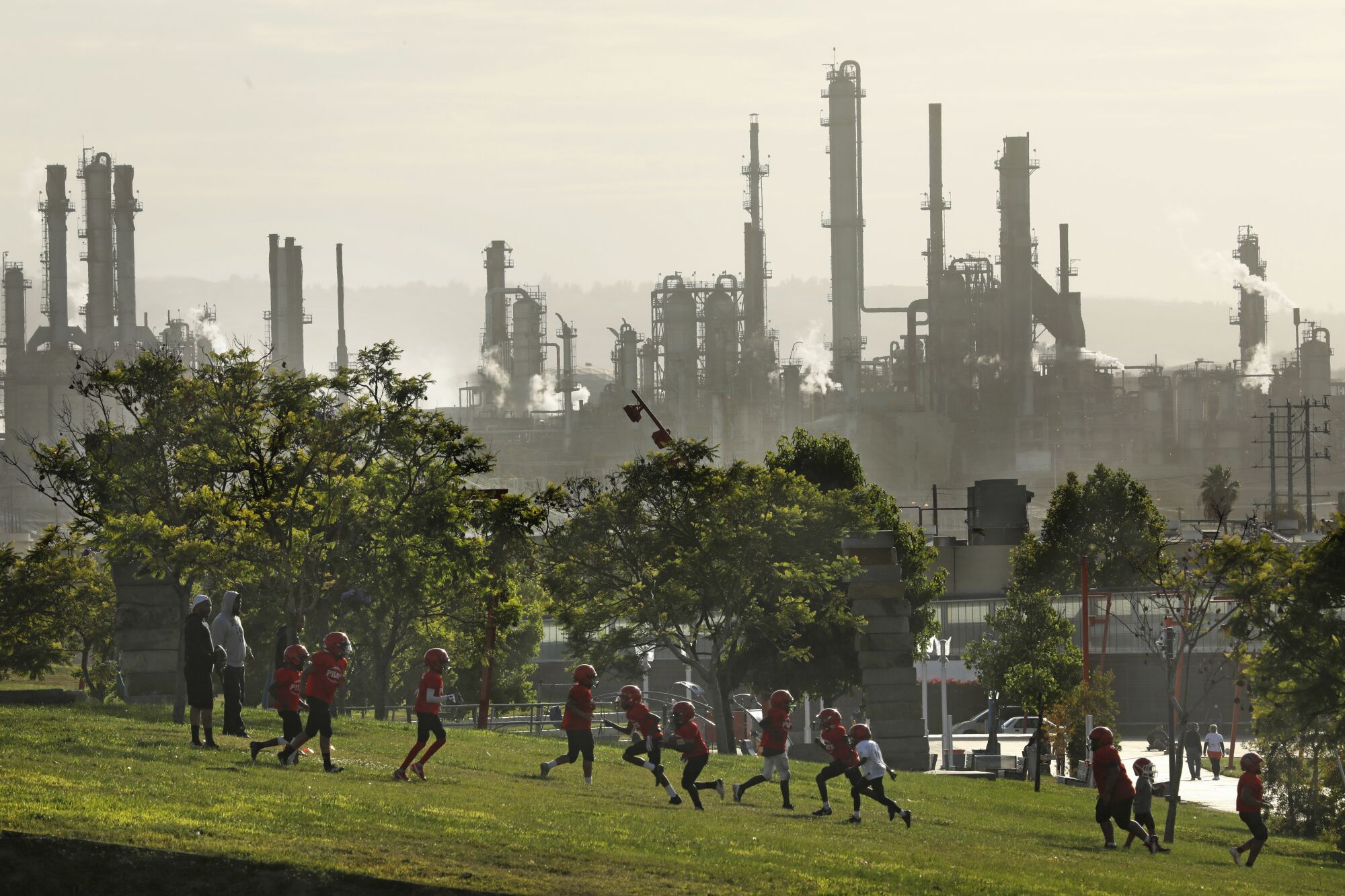 Children in football helmets run in a park with a refinery in the background