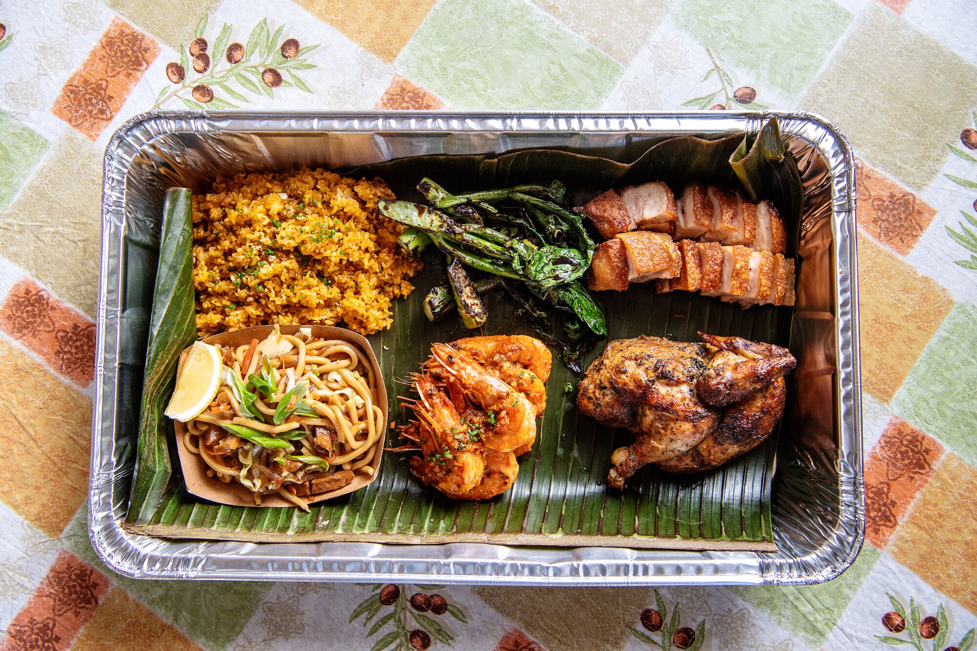 A foil pan contains noodles, prawns, rice, greens and more on a banana leaf.