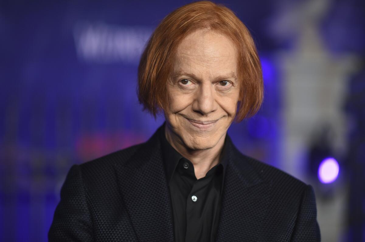 Danny Elfman smiles slyly while clad in a black suit with his red hair in a bob