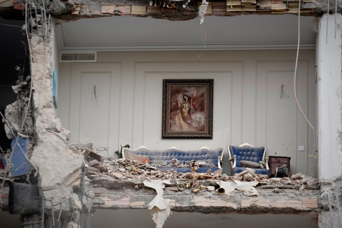 A sheared-off wall exposes furniture covered in debris and a painting on a wall