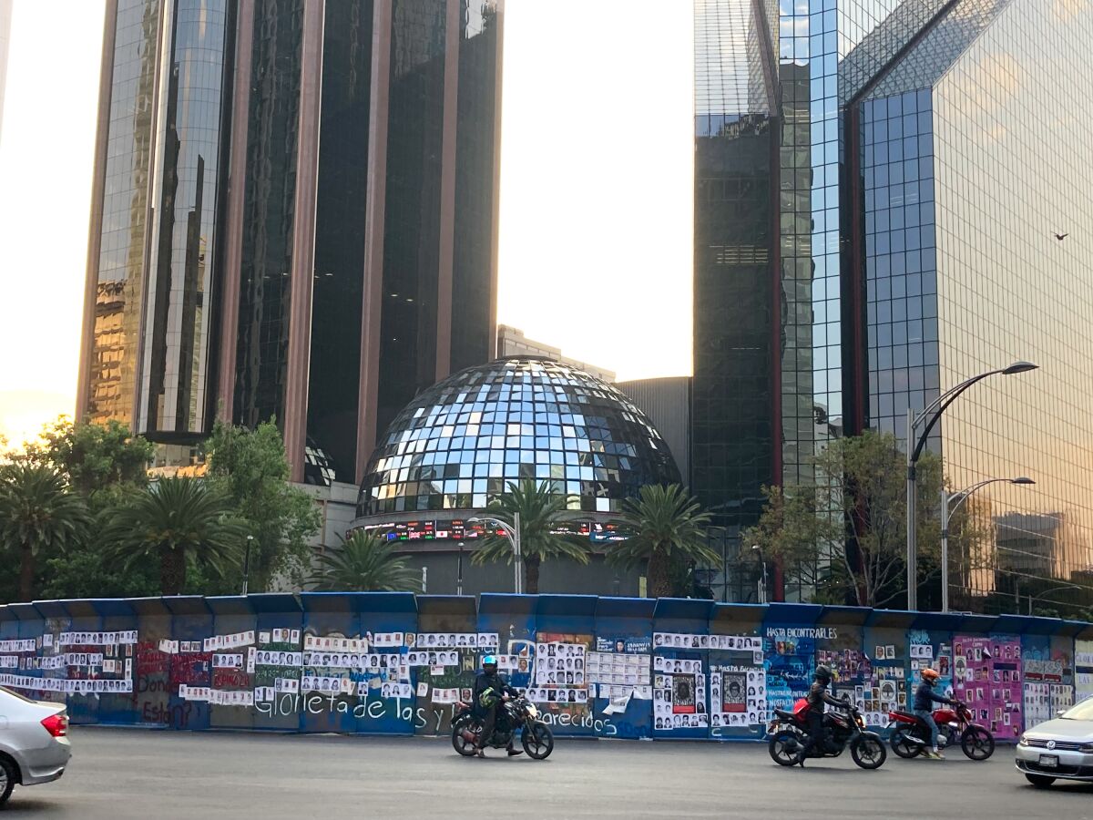 A fence adorned with posters separates motorists from a domed building flanked by two taller buildings