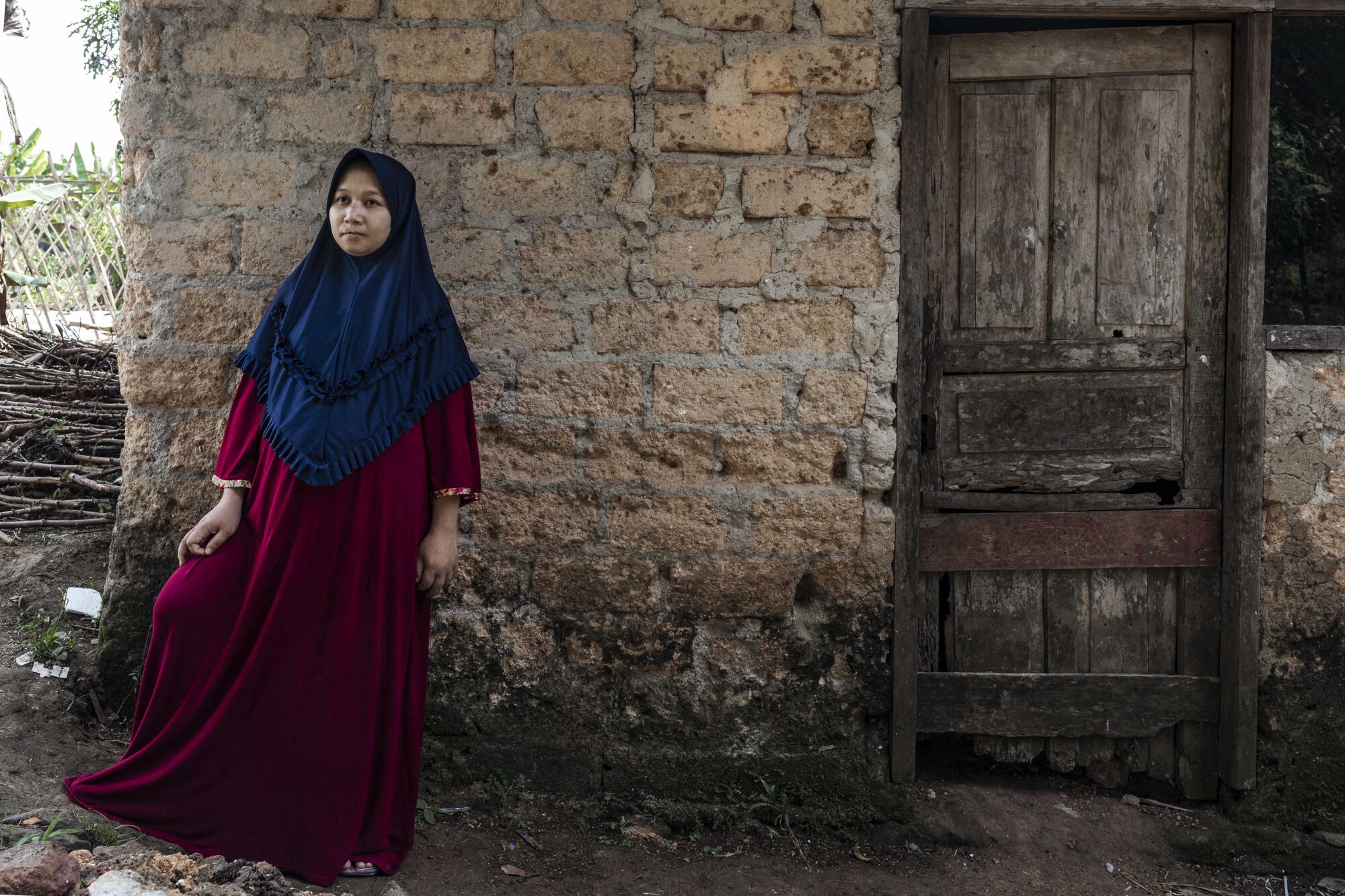 A woman in a red robe and blue hijab stands in front of a brick wall near a wooden door
