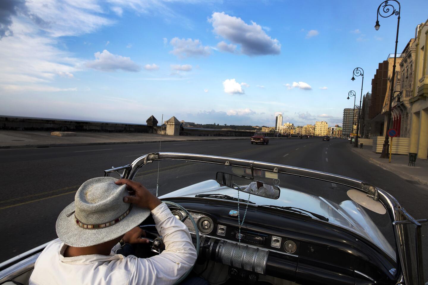 A taxi driver hangs onto his hat while driving his vintage convertible along the Malecon in Havana.