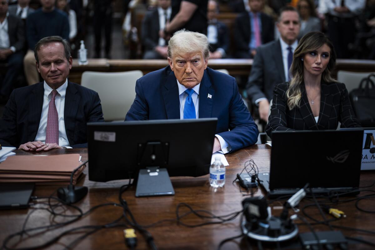 Former President Trump sitting at a table in a courtroom, flanked by two lawyers and with spectators in the background